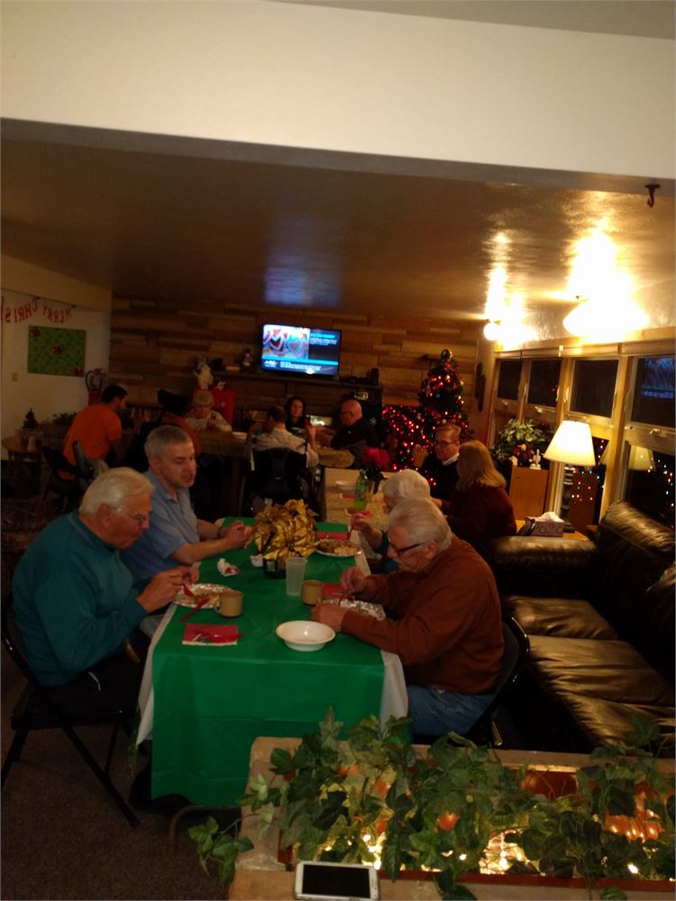 Individuals sharing a Christmas dinner at a long table decorated in green and gold, with a lit Christmas tree behind them