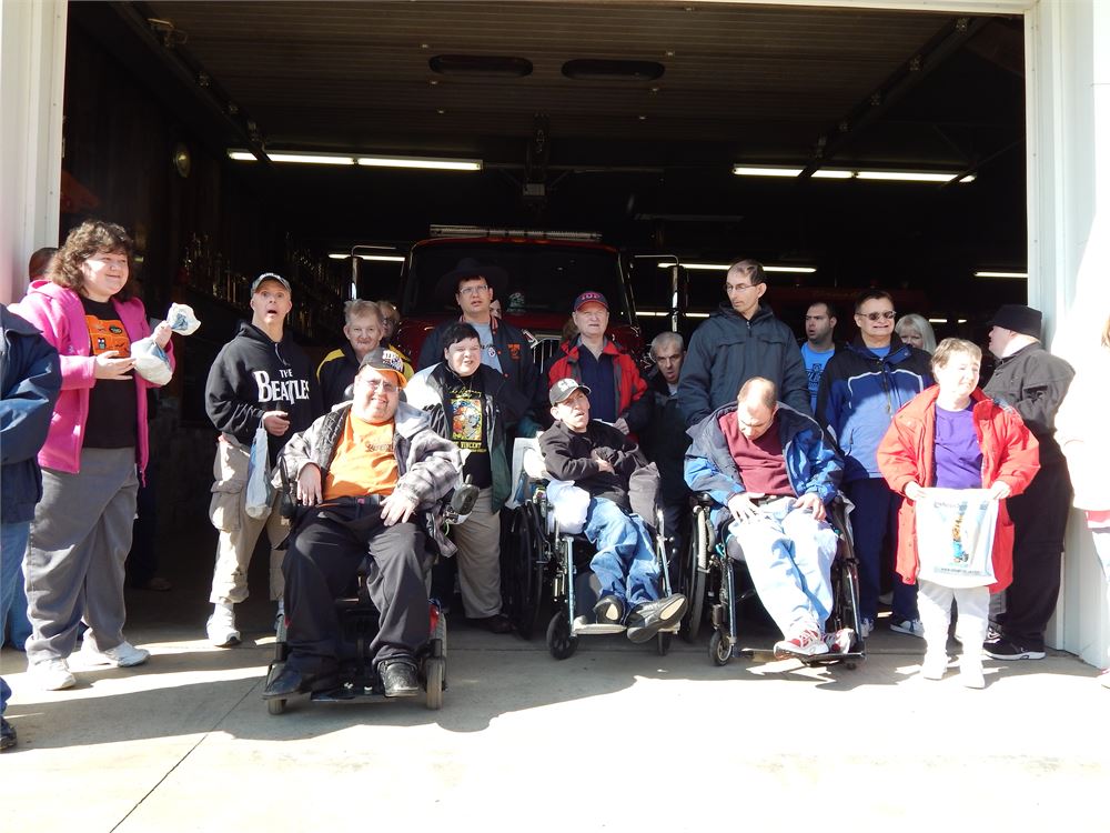A group of individuals and staff gathered in front of a fire engine at the open bay door of a fire station