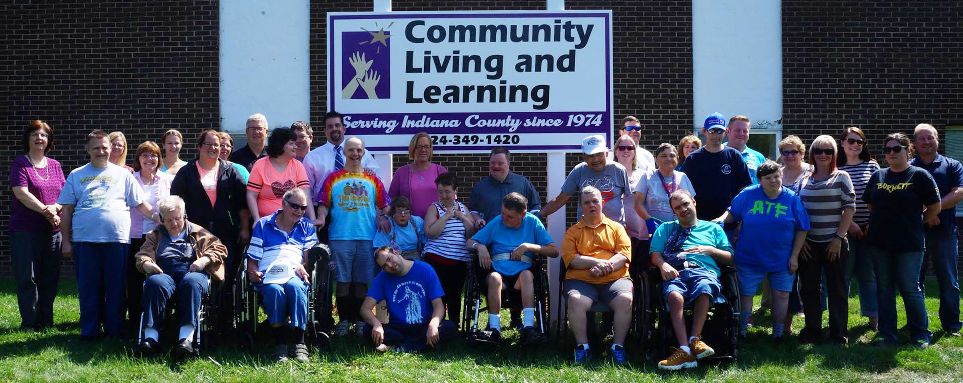 Around forty individuals and staff members of Community Living & Learning gathered together outside the agency's sign on a sunny day