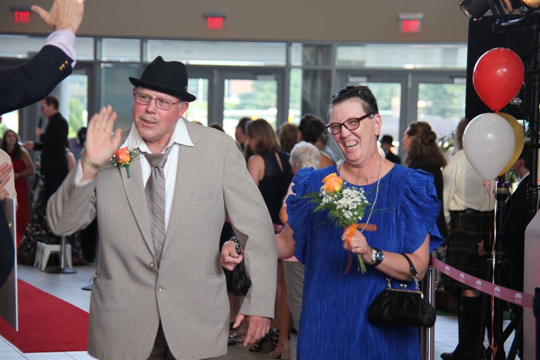 Two individuals walking a red carpet at a formal celebration, wearing corsages and smiling as guests applaud
